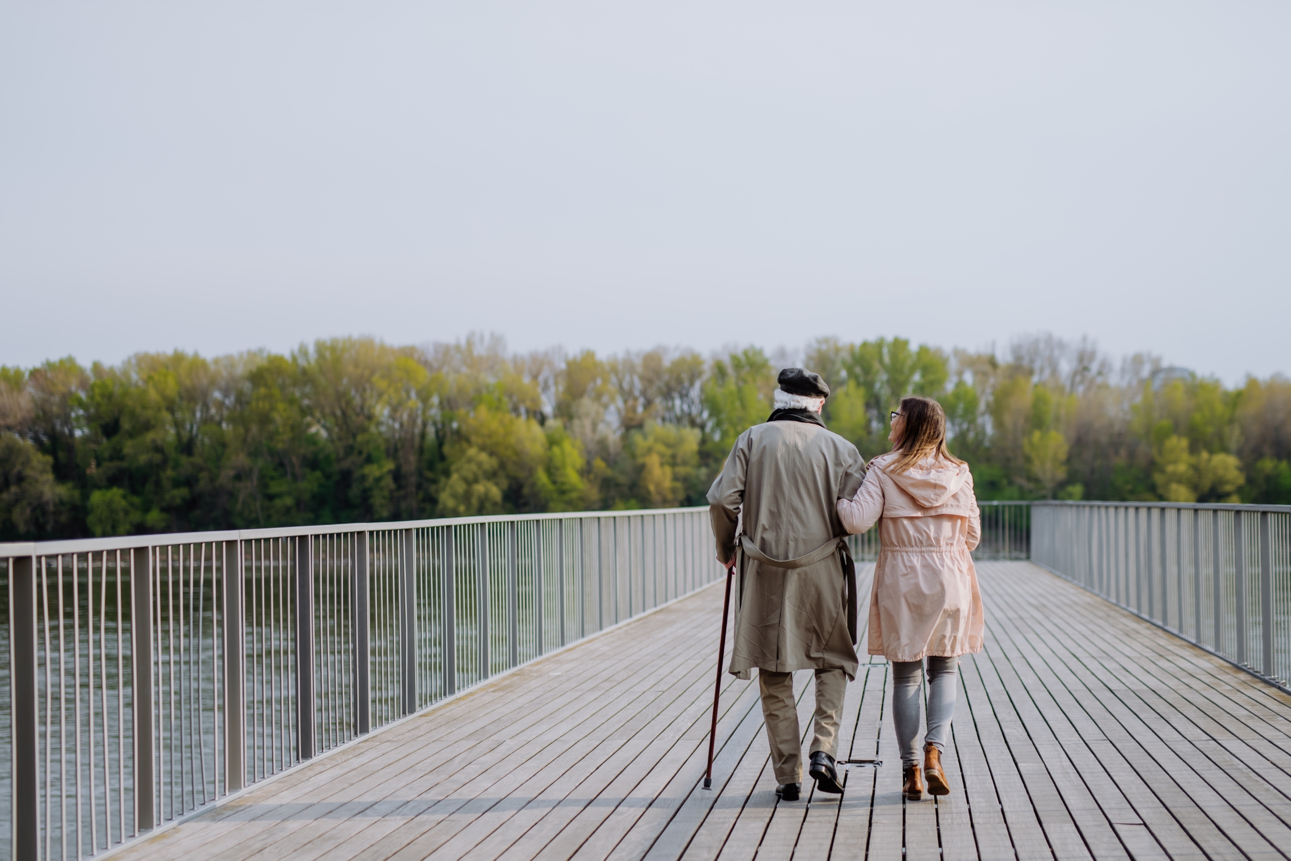 Rear view of senior man with daughter outdoors on a walk on pier by river.